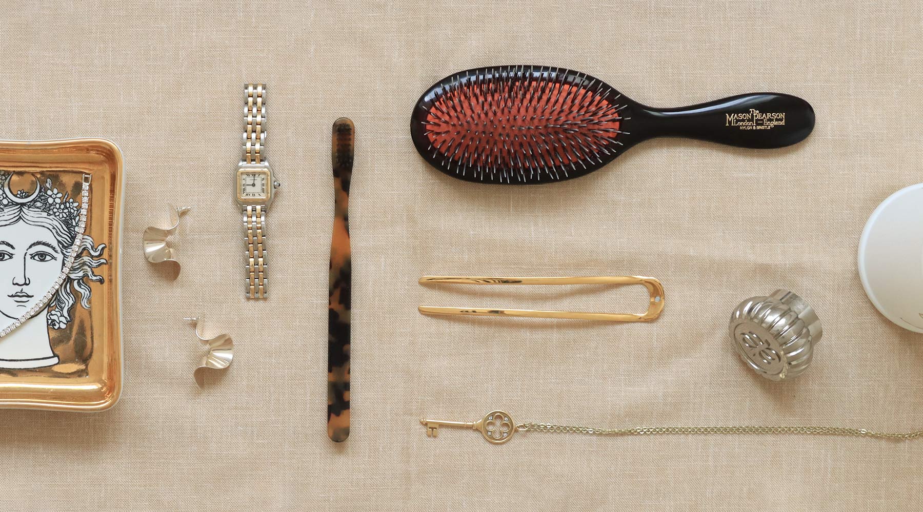 Flat lay of Akla luxury fluoride-free mouthwash cap with Cartier watch, Tiffany & Co necklace, Mason Pearson brush, Fornasetti tray, and high-end toothbrush symbolizing oral wellness as part of luxury lifestyle.