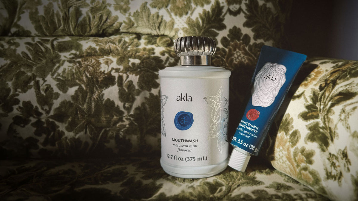 Akla Moroccan Mint Mouthwash and Whitening Toothpaste arranged on a green floral upholstered armchair, featuring the brand’s signature glass bottle and blue aluminum toothpaste tube.