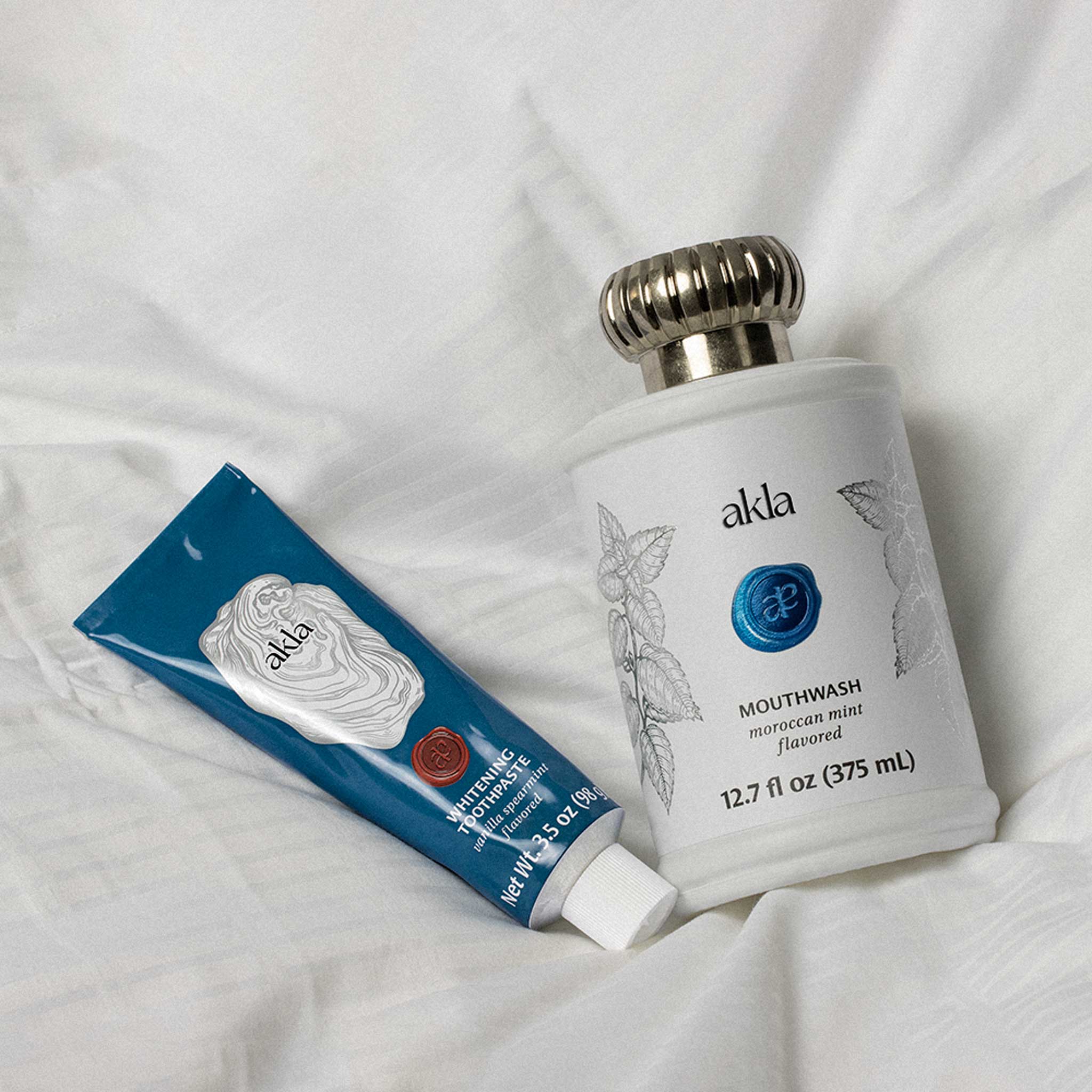 Akla toothpaste tube in blue aluminum tube and mouthwash in glass bottle on a bed, morning and night self care rituals. 