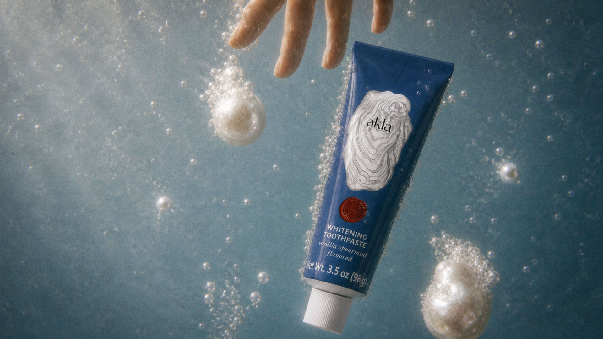 Akla's luxury ora care toothpaste underwater with pearls and a hand reaching for the tube. 