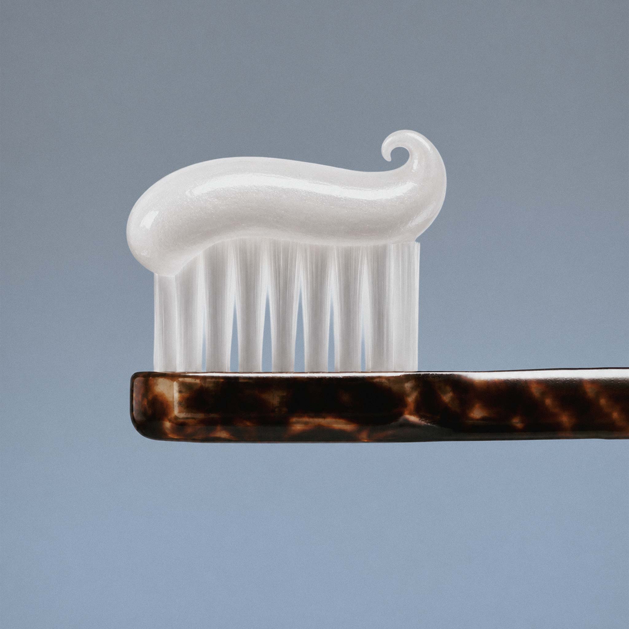 A close-up of toothbrush with dark brown tortoiseshell handle and white bristles, topped with Akla hydroxyapatite toothpaste and set against a blue-gray background.