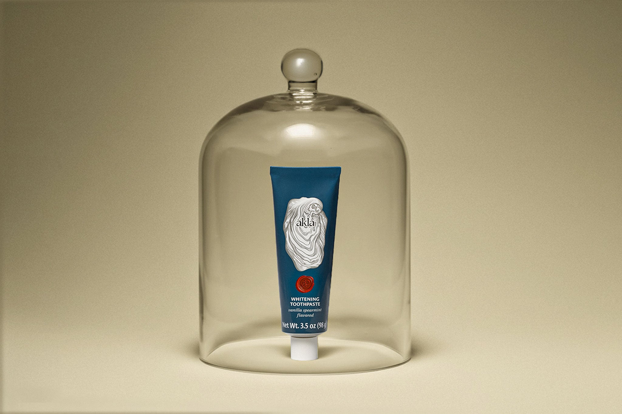 Akla toothpaste tube under a glass apothecary dome on a muted yellow background.