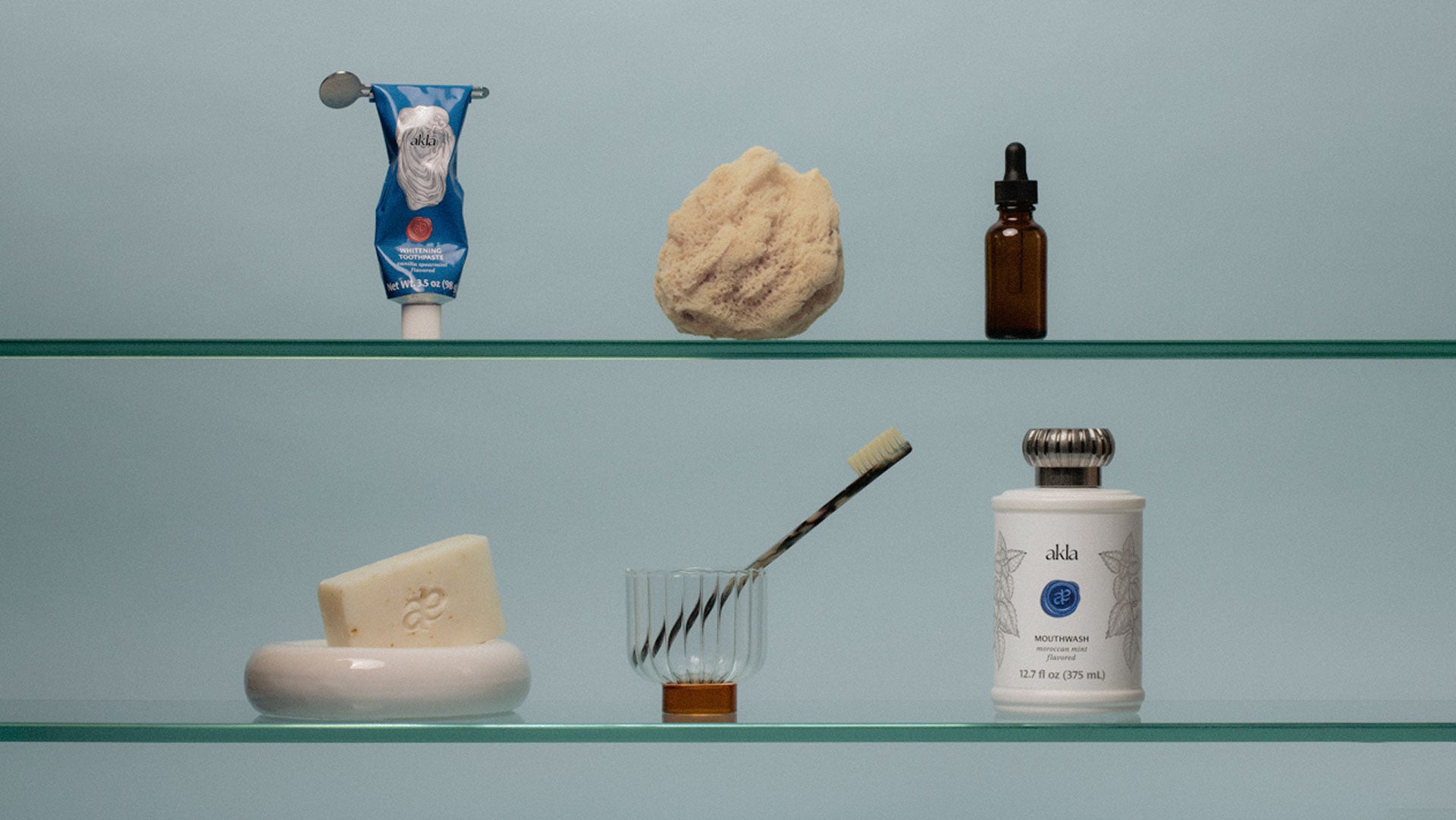 Akla luxury oral care and collection of self care products on a glass shelf against a light blue background.