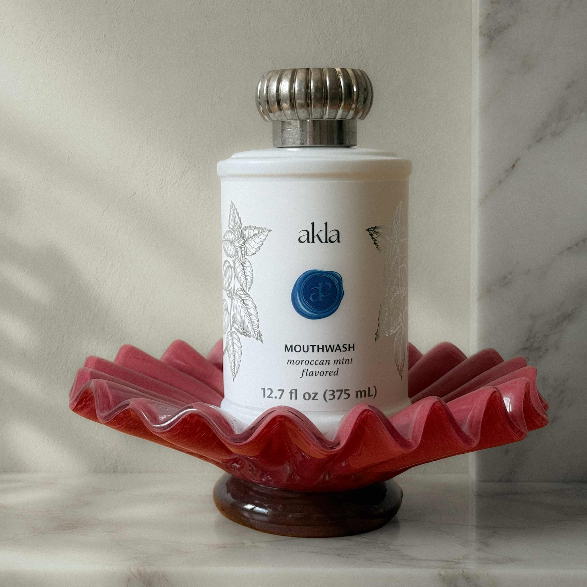 Akla alcohol-free mouthwash in a white glass bottle displayed on a sculptural glass dish — Swiss-made luxury oral care.