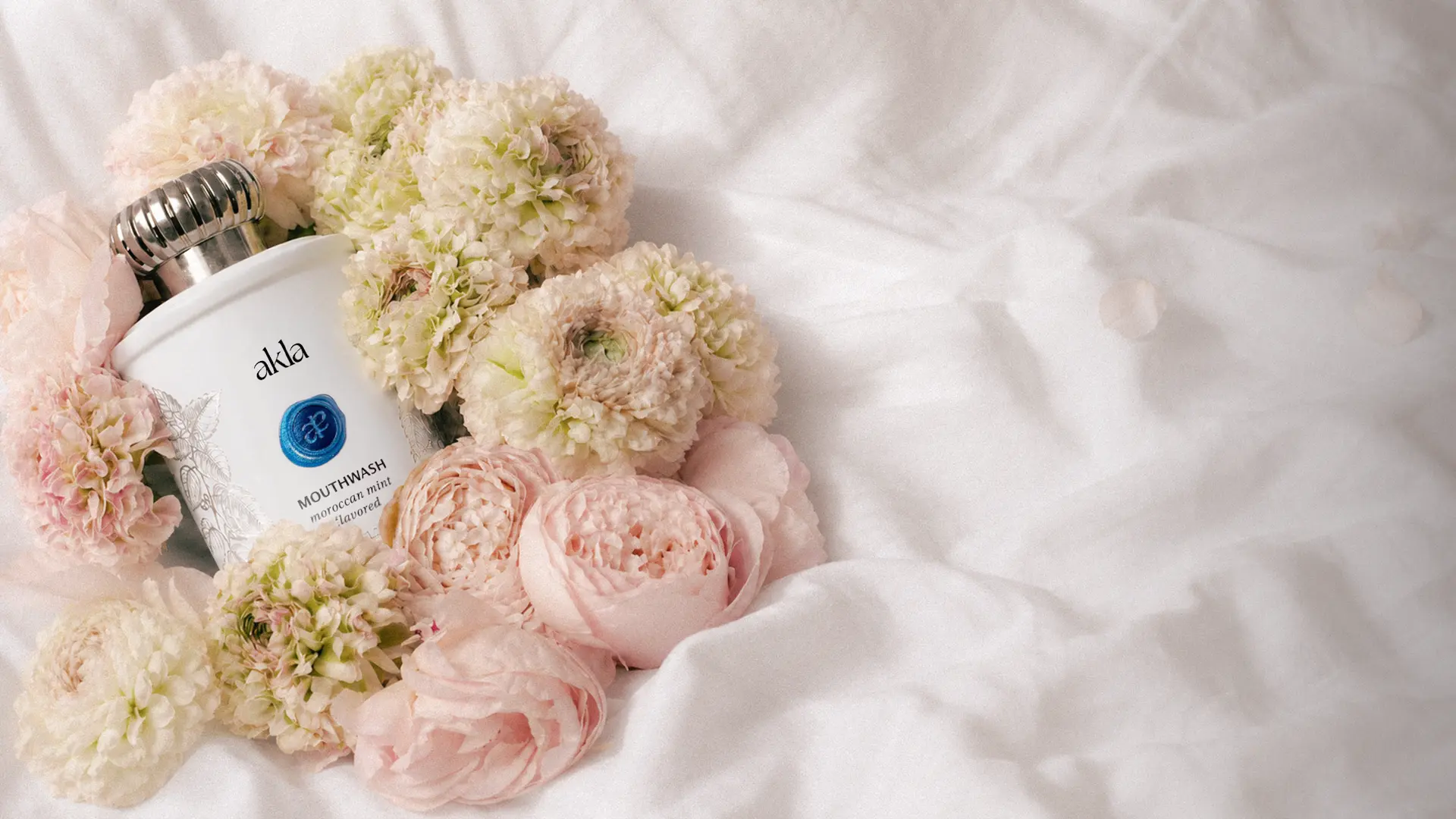 Akla Moroccan mint mouthwash in a silver-capped glass bottle resting among pale pink and cream garden roses on white linen.