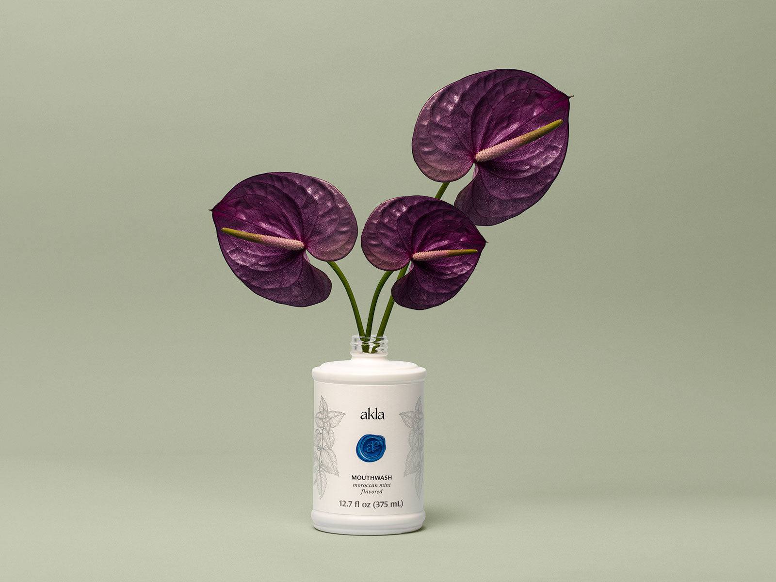 Akla Moroccan Mint Mouthwash bottle styled as a vase with two deep purple anthurium flowers against a soft green background.
