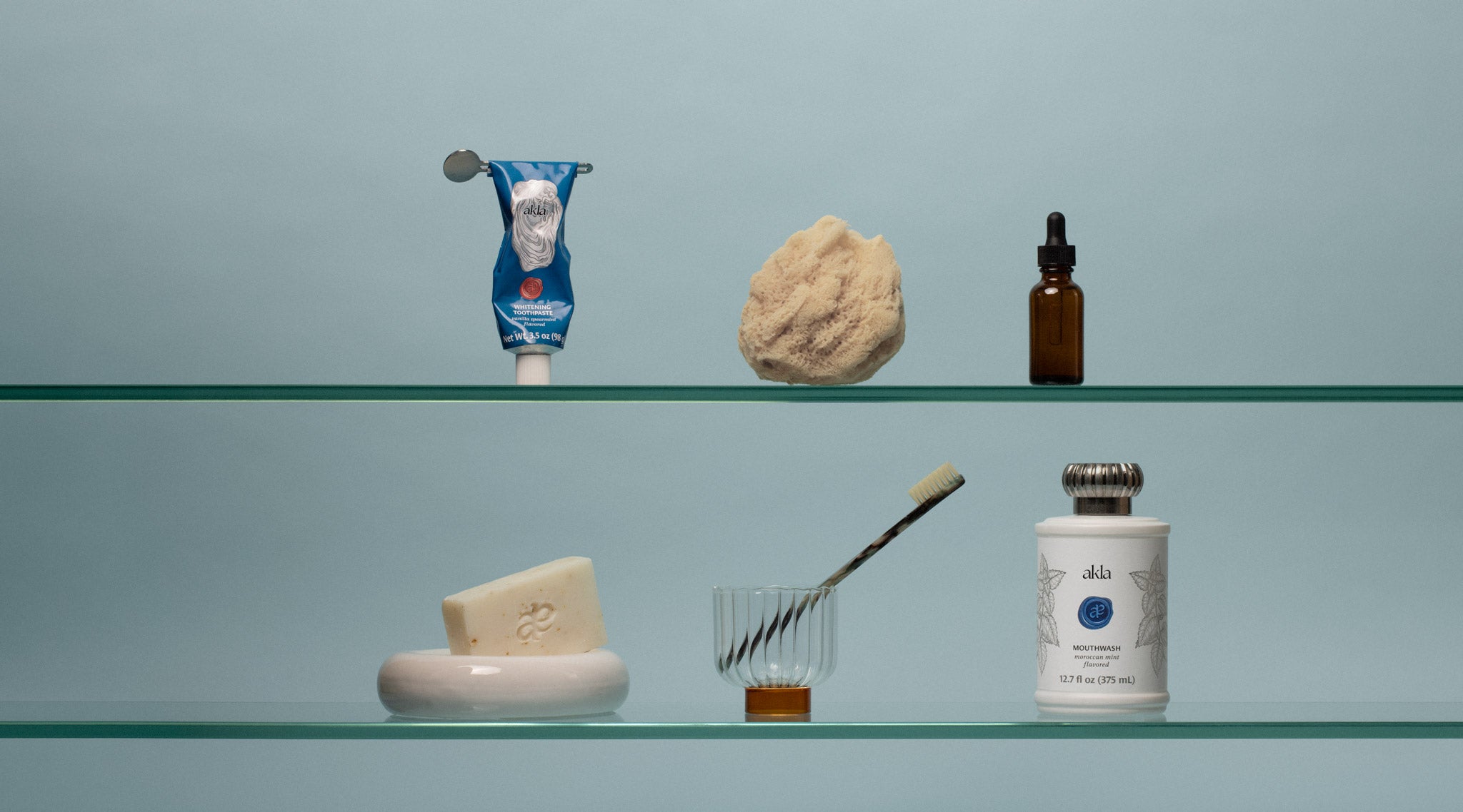 Fluoride-free luxury toothpaste and mouthwash by Akla on a glass shelf, with toothbrush, sponge, soap, and dropper bottle.